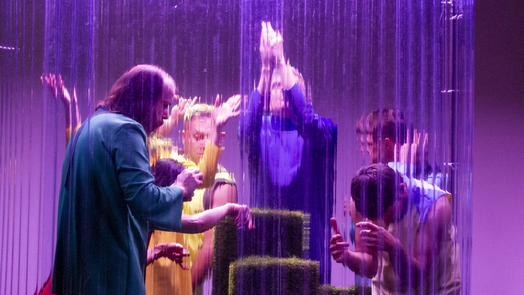Szenenfoto aus einer Auffuehrung: Mehrere Personen hinter einer Art Glasscheibe auf der Bühne.
