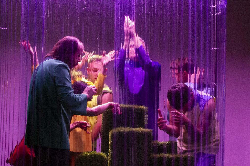 Szenenfoto aus einer Auffuehrung: Mehrere Personen hinter einer Art Glasscheibe auf der Bühne.