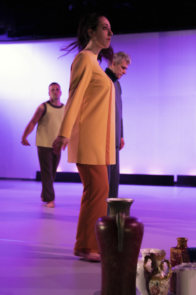 Scene photo of a performance: vases in the foreground. In the background three people in a stage.