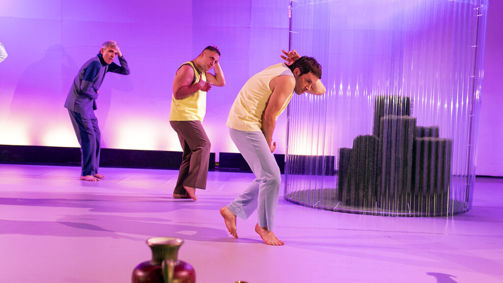 Scene photo of a performance: a couple people behind a glassScene photo of a performance: Three men stand on a stage. In the background is a green house.