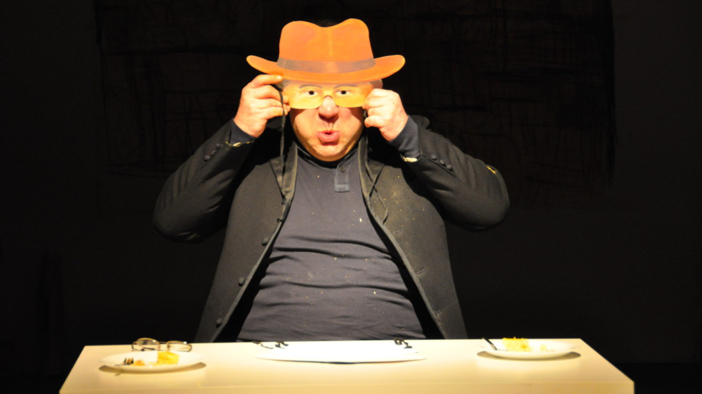 Szenenfoto aus einer Auffuehrung: Ein Mann sitzt an einem Tisch auf einer Theaterbühne und hält sich eine Maske vor das Gesicht.