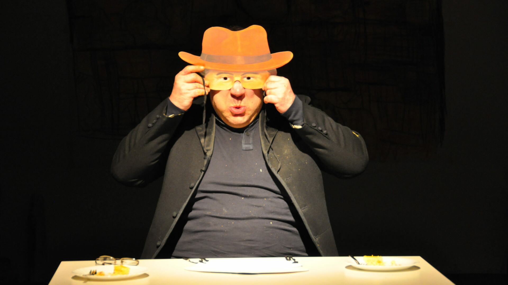 Photo from a performance: A man sits at a table on a theater stage and holds a mask in front of his face.