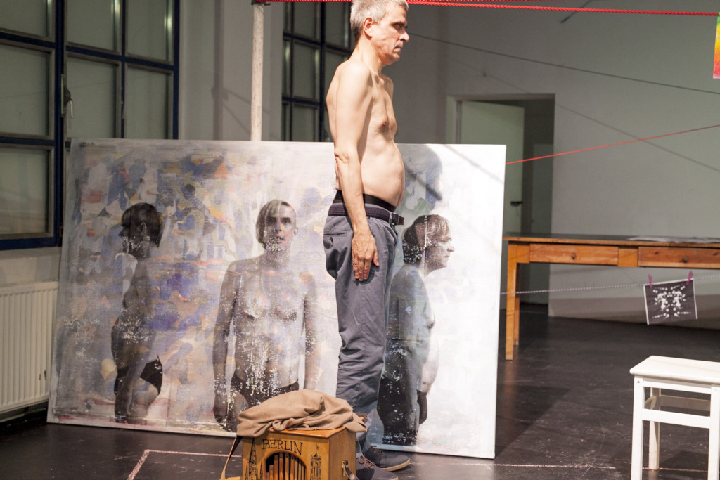 Szenenfoto aus einer Auffuehrung: Ein Mann mit geschlossenen Augen steht auf einer Theaterbühne vor einer Leinwand.