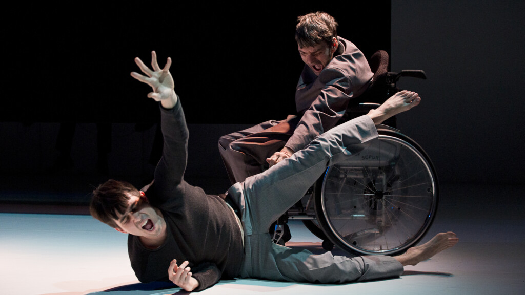 Scene photo of a performance: One men in a wheelchair on a stage. Another one is laying on the ground in front of him.