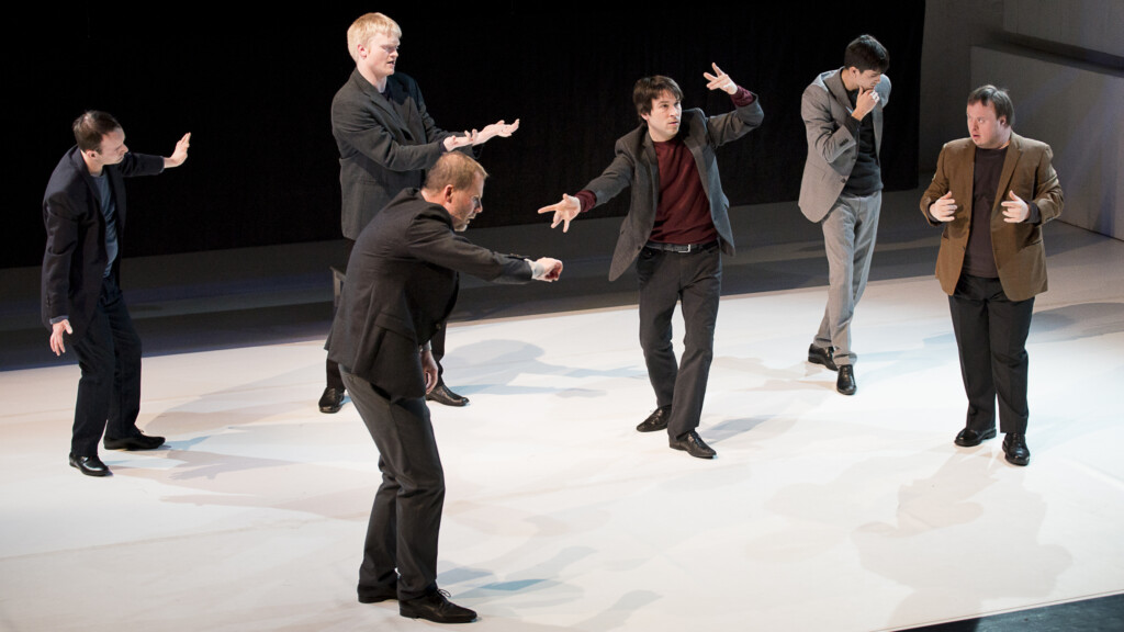 Scene photo of a performance: Six men in a suit on a stage.