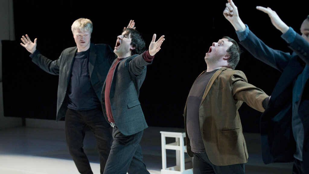 Scene photo of a performance: Four men in suits with wide-open arms on a stage.