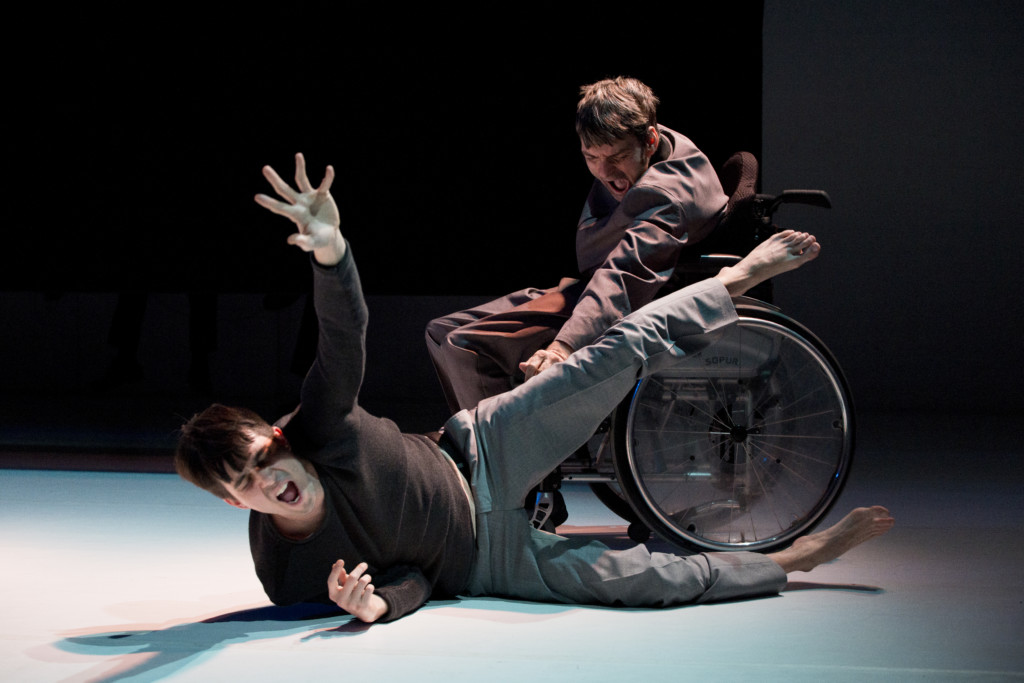 Scene photo of a performance: Two men are screaming. One is lying on the ground. The other one is in a wheelchair.