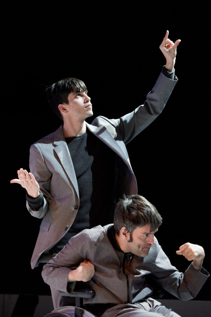 Scene photo of a performance: Two men with tilted arms on a stage. One in a wheelchair. The other one behind him.