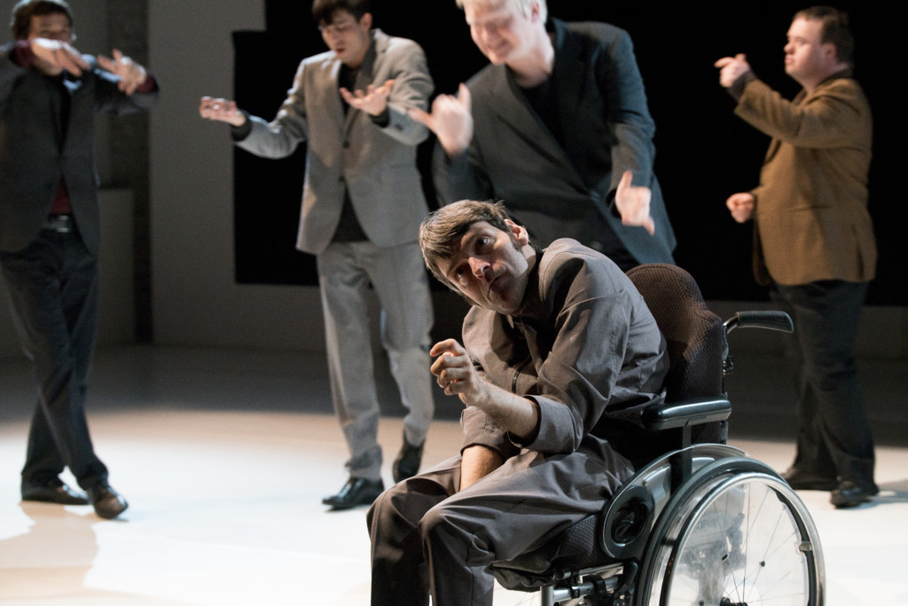 Scene photo of a performance: Five men on a stage. One is in a wheelchair. The other four blurred in the background.