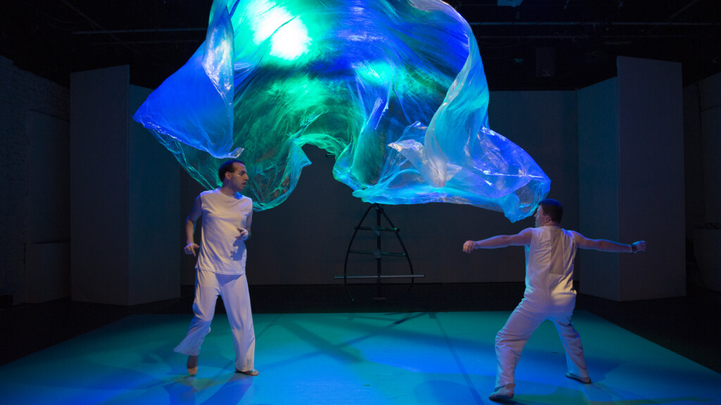 Scene photo of a performance: Two men on a stage. A plastic foil between them.