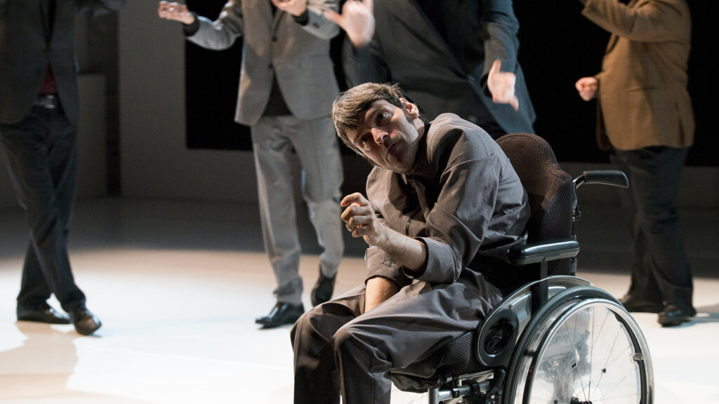 Scene photo of a performance: Five men on a stage. One is in a wheelchair. The other four blurred in the background.