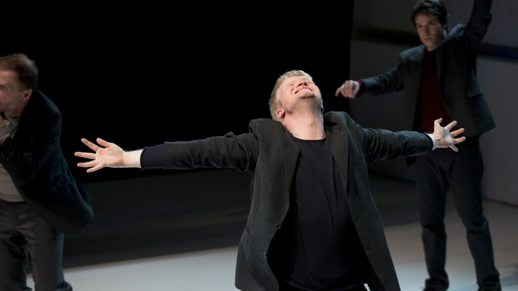 Scene photo of a performance: Three men on a stage. One men in the foreground with wide open arms. Two other men in the background.