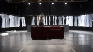 Szenenfoto aus einer Auffuehrung: Zwei Sessel in der Mitte. Ringsherum Kleider auf einer Kleiderstange.