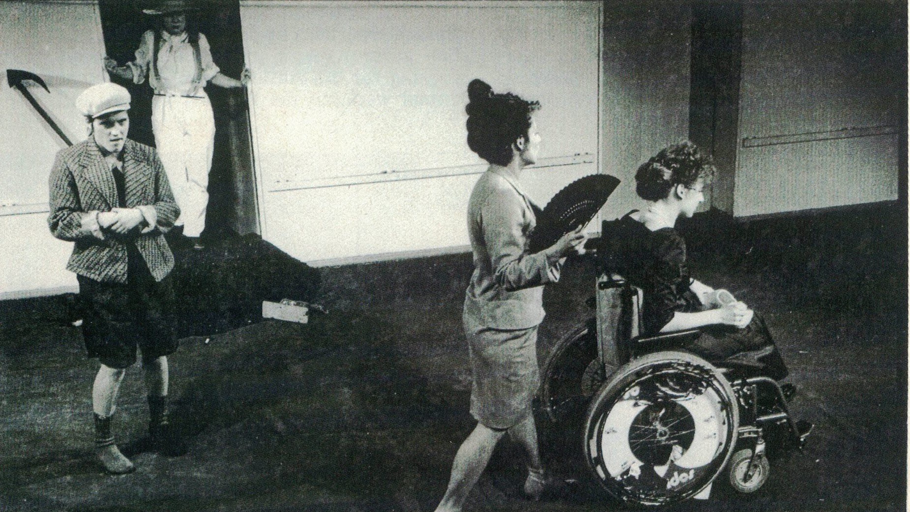 Szenenfoto aus einer Auffuehrung: Vier Menschen auf der Bühne. Im Vordergrund schiebt eine Frau mit Fächer eine andere Frau im Rollstuhl. Links daneben steht ein Junge mit fragender Geste. Im Hintergrund kommt jemand mit Strohhut aus einer Tür.