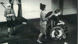 Szenenfoto aus einer Auffuehrung: Vier Menschen auf der Bühne. Im Vordergrund schiebt eine Frau mit Fächer eine andere Frau im Rollstuhl. Links daneben steht ein Junge mit fragender Geste. Im Hintergrund kommt jemand mit Strohhut aus einer Tür.