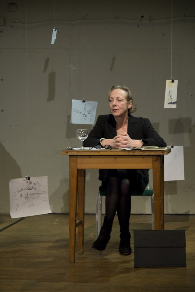 Scene photo of a performance: One woman is sitting on a wood table.