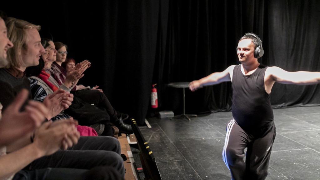 Szenenfoto einer Auffuehrung: Mann auf einer Theaterbuehne mit Kopfhörern. Vor Ihm das Publikum.