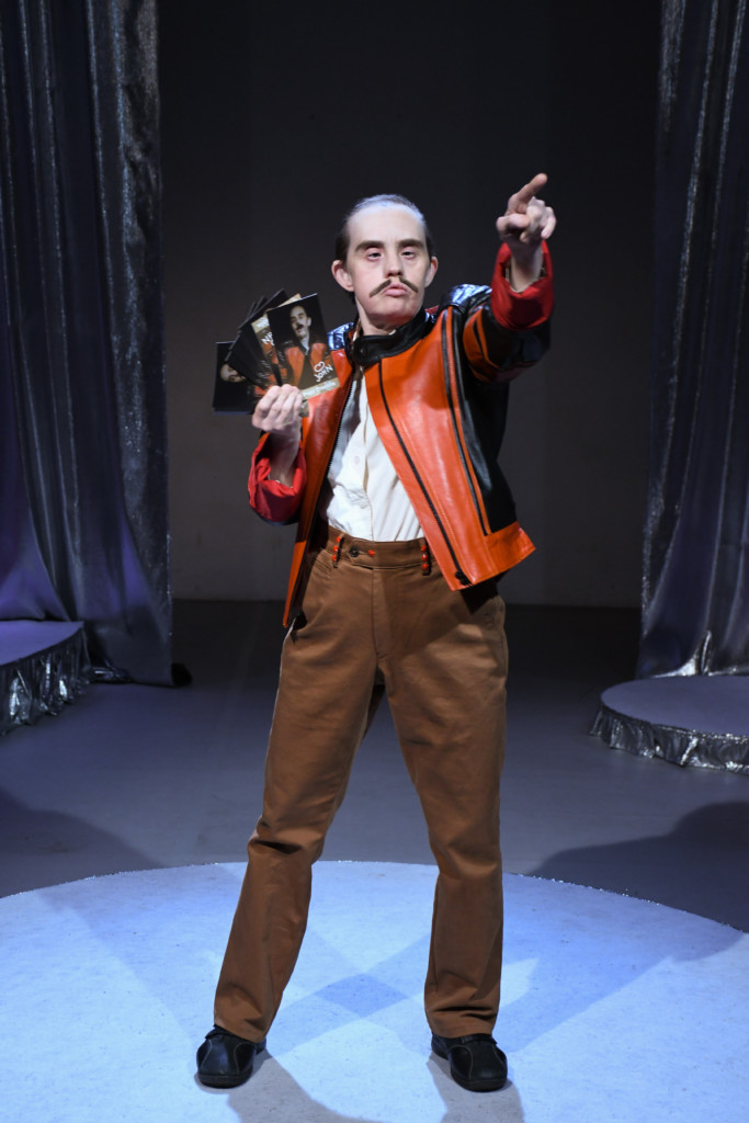 Szenenfoto einer Auffuehrung: Eine Person steht auf einer Bühne mit drei Karten in der linken Hand und zeigt auf etwas mit der rechten Hand.