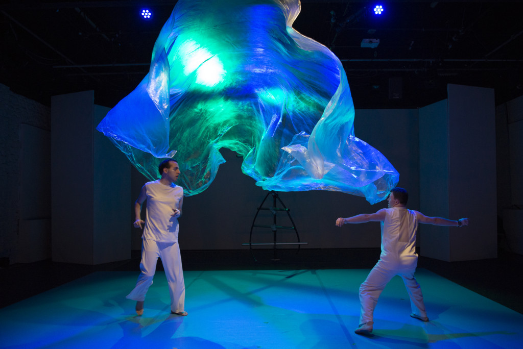 Scene photo of a performance: Two men on a stage. A plastic foil between them.