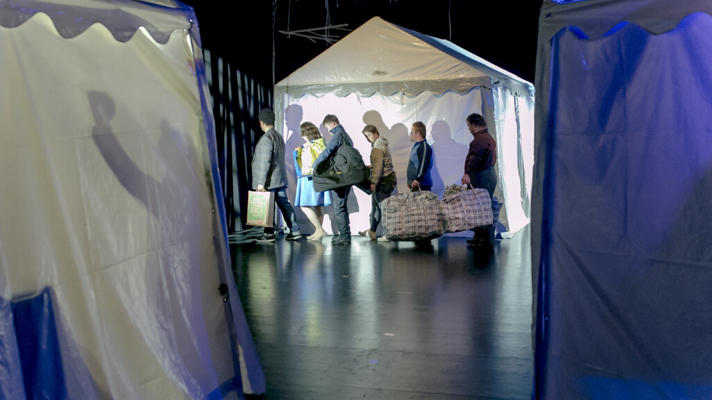 Scene photo of a performance: Three white party tents, 6 people mit bags in the background.