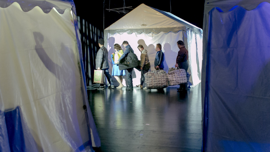 Szenenfoto aus einer Auffuehrung: Drei weisse Partyzelte, zwei angeschnitten im Vordergrund, eines im Hintergrund. Vor dem hintern Zelt geht eine Gruppe von 6 Personen hintereinander entlang, in Alltagskleidung, alle mit großen Tüten und Taschen.