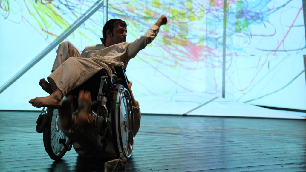 Scene photo of a performance: A man in a wheelchair in front of something which looks like map an a stage.