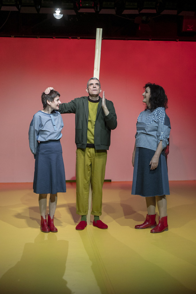 Scene photo of a performance: One man in the middle.Behind him a piece of wood. A woman to the left and to the right side.