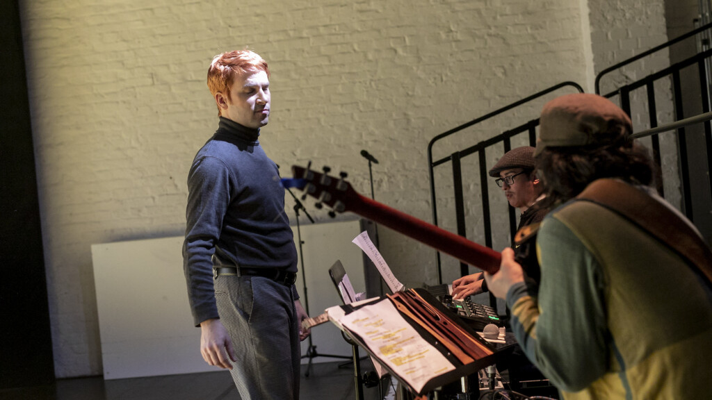 Scene photo of a performance: There men on a stage. Two are playing an instrument. One is standing.