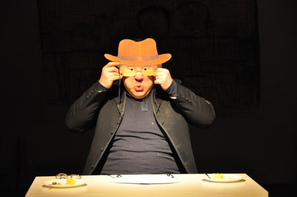 Szenenfoto aus einer Auffuehrung: Ein Mann sitzt an einem Tisch auf einer Theaterbuehne und hält sich eine Maske vor das Gesicht.