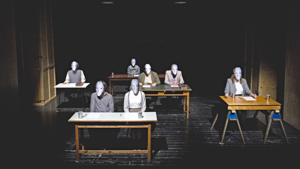 Five tables are on the stage. At every table there is a person sitting wearing modest clothes and a grey mask. At the table in the middle there are two people sitting.