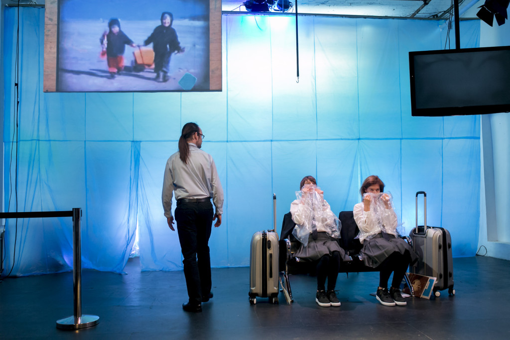 Scene photo of a performance: Three people on a stage. Two women are sitting and one man is standing.