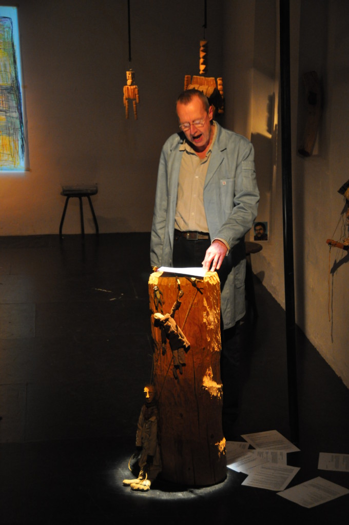 Photo from a performance: One man is reading something from a paper. wood figures are hanging behind him.