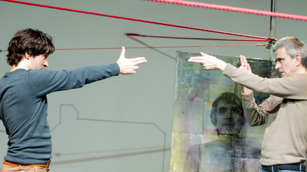 Scene photo of a performance: Two men stand in front of each other an a stage pointing at each other.
