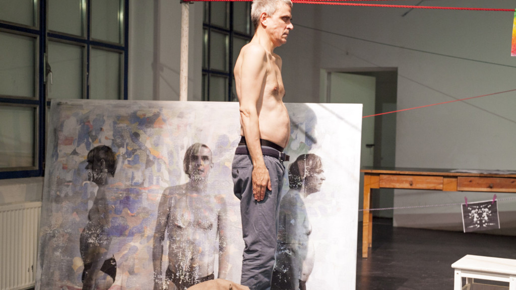 Szenenfoto aus einer Auffuehrung: Ein Mann mit geschlossenen Augen steht auf einer Theaterbühne vor einer Leinwand.