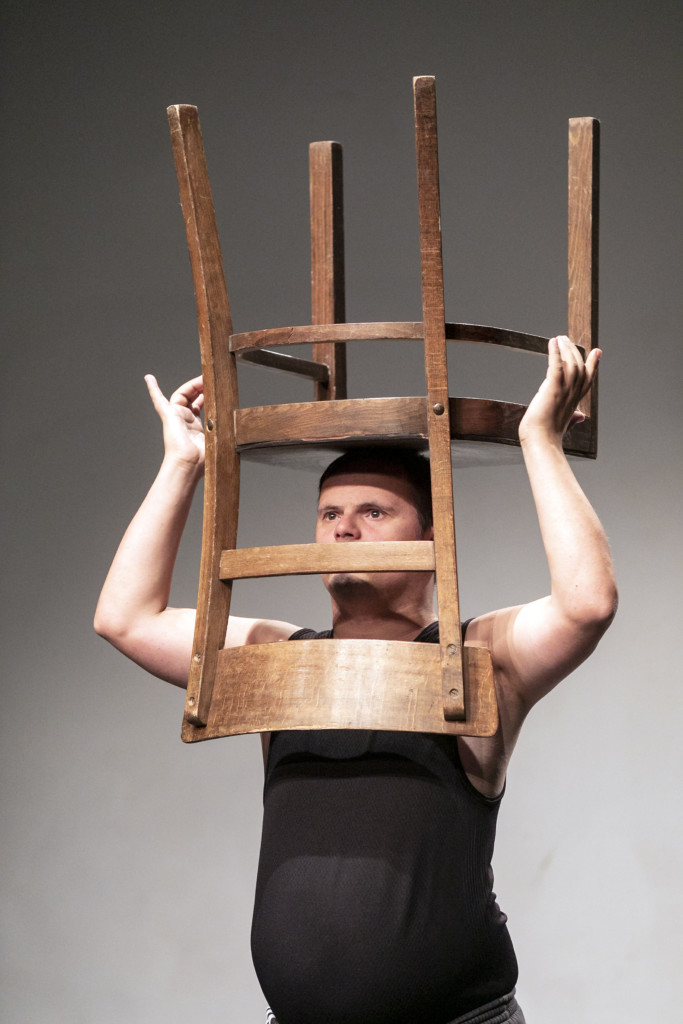 Scene photo of a performance: A man holding a chair over his head on a stage.