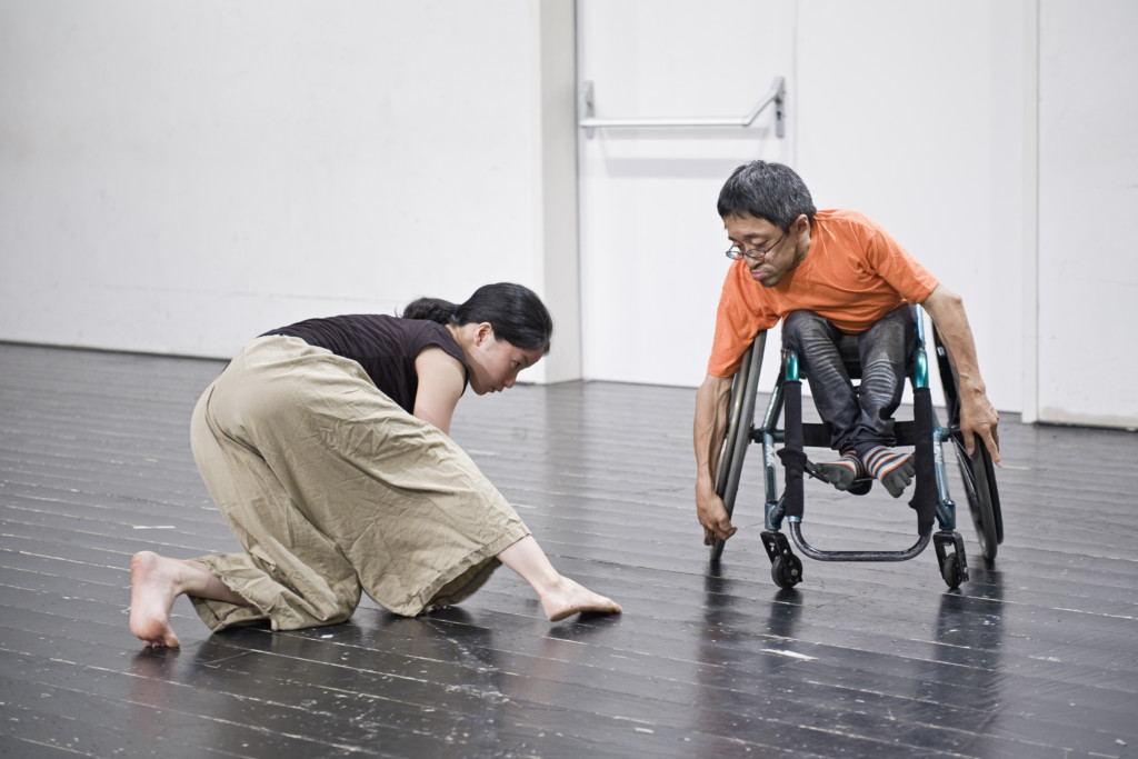 Szenenfoto: Ein Mann im Rollstuhl und eine Frau auf Knieen in Bewegung. Beide sind japanisch