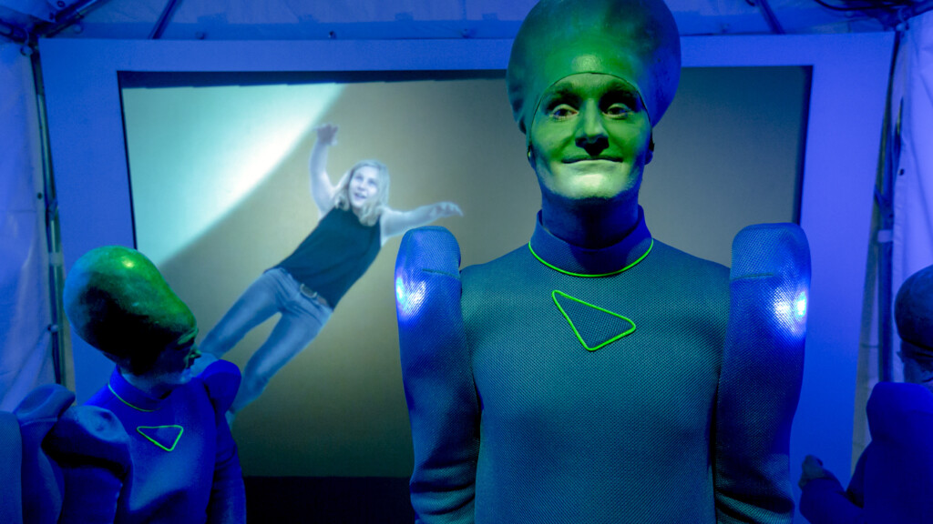 Szenenfoto aus einer Aufführung: Ein futuristisch gekleideter Mann mit einen verlängerten Hinterkopf lächelt in die Kamera.