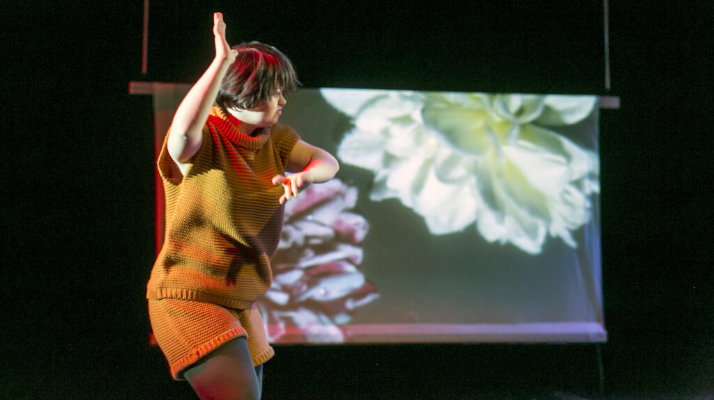 Szene aus einer Aufführung: Eine Frau im gelben Strickkleid tanzt vor einer Leinwand auf der große Blüten zu sehen sind