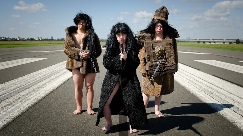 Three people in archaic fur coats on an airfield. The woman in the middle holds a gun.