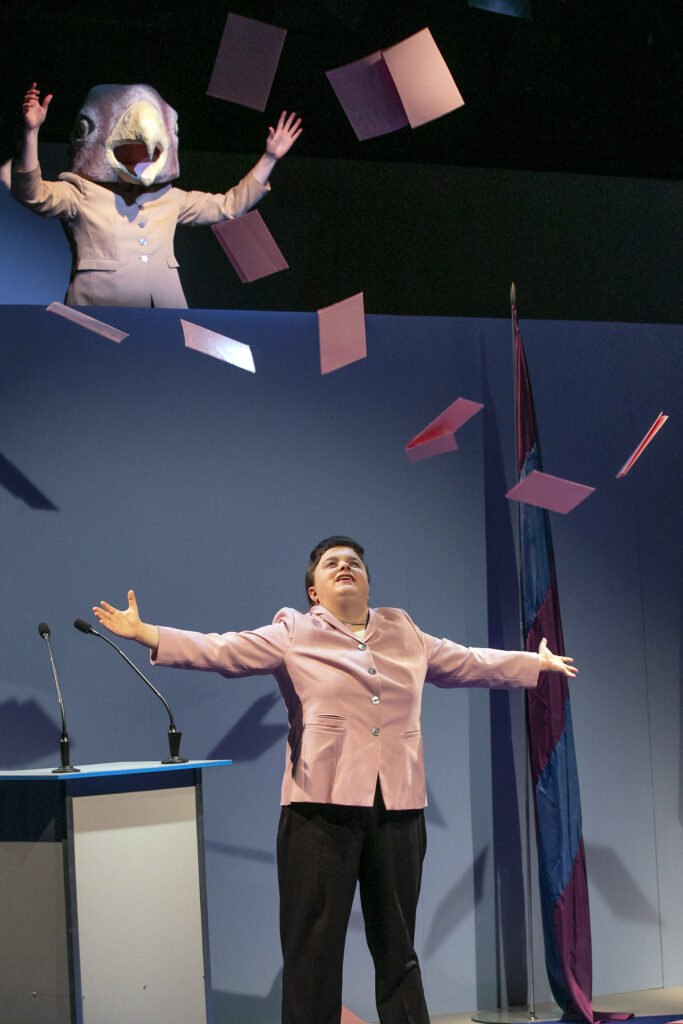 Scene photo of a performance: Upstairs a person mit eagle mask. downstairs a woman with wide spread arms. It is raining ballot paper.