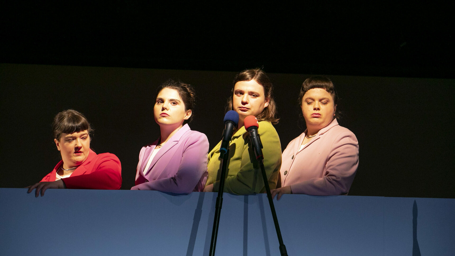 Szenenfoto einer Auffuehrung: Vier Frauen einer einer Bühne in Blazern hintereinander.