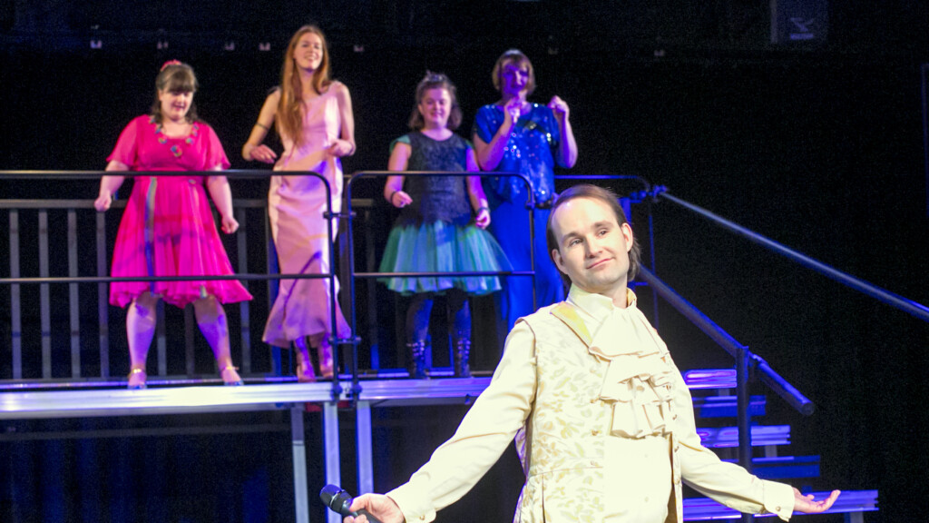Scene photo of a performance: A person with wide-open arms in the foreground. Four other people in the background.