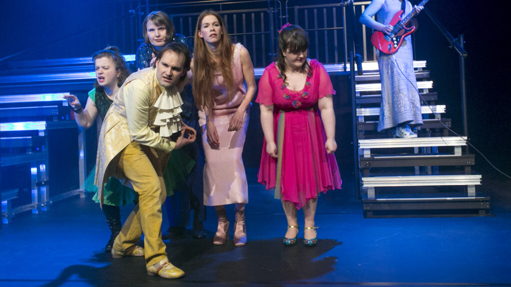 Scene photo of a performance: Four people on a stage. In the background a person with a guitar.