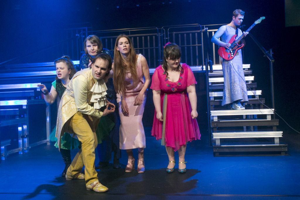 Scene photo of a performance: Four people on a stage. In the background a person with a guitar.