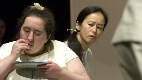 Szenenfoto aus einer Aufführung: Zwei Frauen. Eine isst etwas von einem Teller. Die andere beobachtet sie dabei