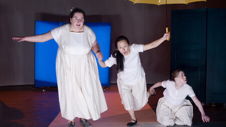 Scene photo of a performance: Three women on a stage. One with a yellow umbrella.