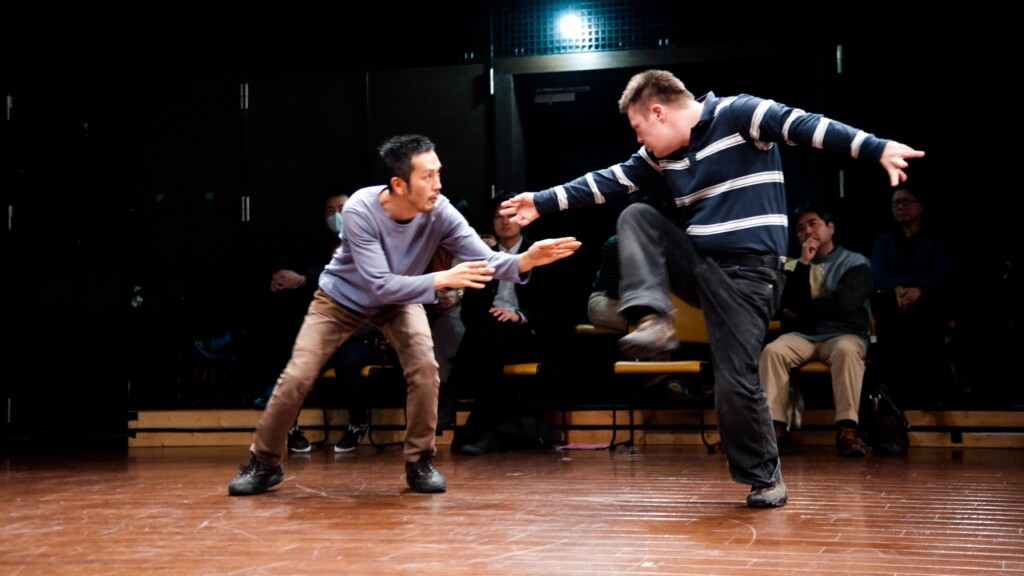 Szenenfoto einer Aufführung: Zwei Männer in Alltagskleidung tanzen.