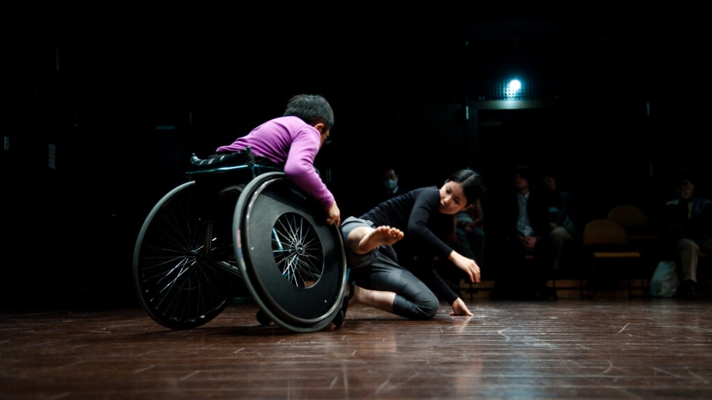 Szenenfoto aus einer Aufführung: Ein Mann im Rollstuhl und eine junge Frau tanzen akrobatisch