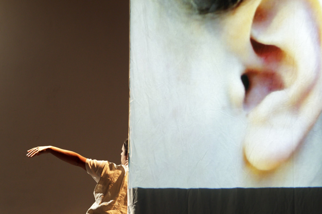 A huge projection of a human ear. In front you see a male dancer, only half of his body shown in the picture.