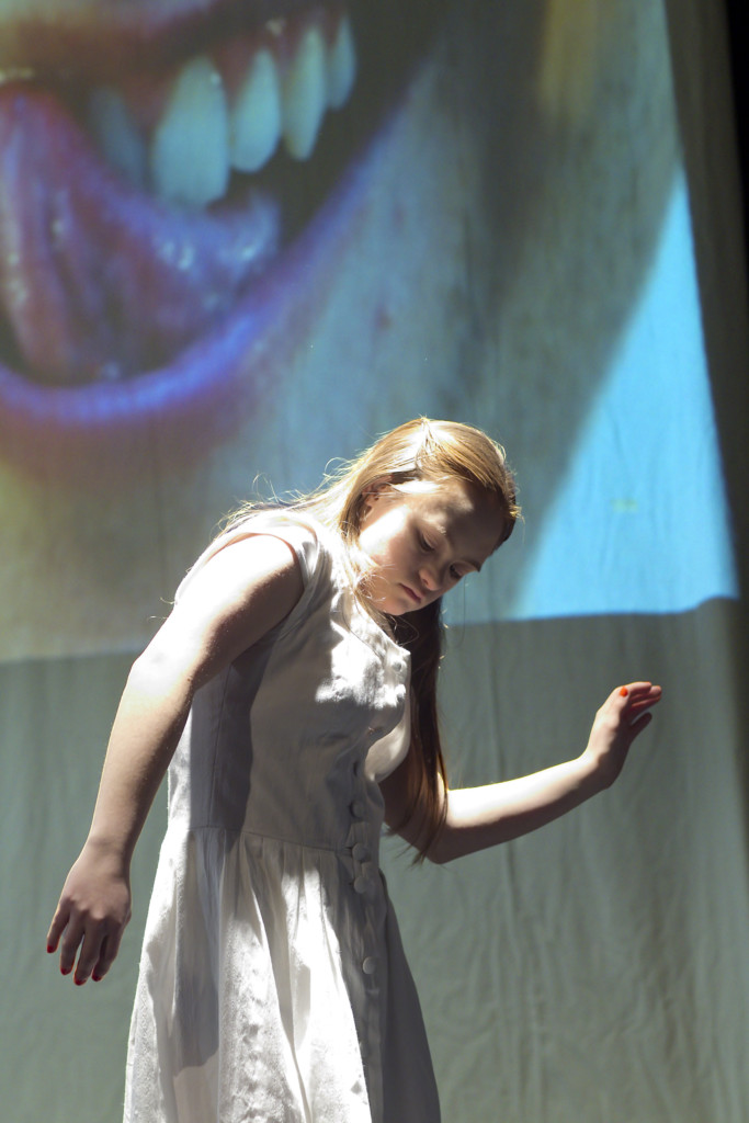 A young female dancer in a white dress moving in front of a visual of a mouth with a stretched out tongue.